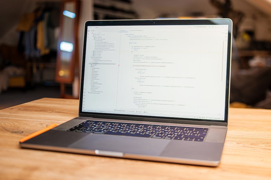A close-up view of a modern silver laptop placed on a wooden desk in a well-lit room. The laptop screen displays lines of code in a text editor, with the interface featuring a sidebar of folders and a
